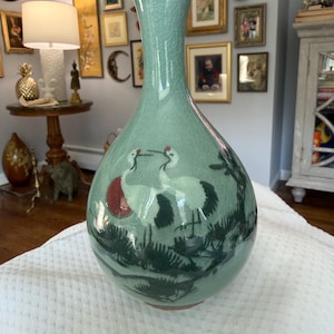 May include: A celadon ceramic vase with a long neck and a rounded body. The vase has a hand-painted design of two cranes with red accents and dark green foliage. The vase is approximately 30 cm tall.