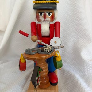May include: A wooden nutcracker figure wearing a black hat with yellow trim, a red jacket, and blue trousers. The figure stands at a round wooden table, holding a silver tool and a circular saw blade, with small hanging ornaments attached to the table.