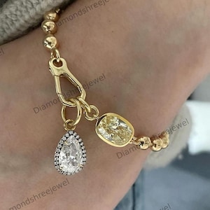May include: A gold bracelet featuring two diamond pendants. One is a pear-shaped diamond with a halo of smaller diamonds, the other is an oval yellow diamond. The bracelet has a gold clasp and a beaded chain.
