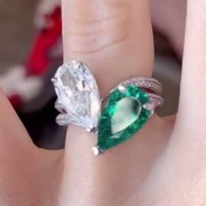 Puede incluir: Un anillo de dos piedras con una gema verde en forma de pera y una gema transparente en forma de pera. El anillo tiene una banda plateada con pequeñas piedras de acento. El anillo se lleva en un dedo.