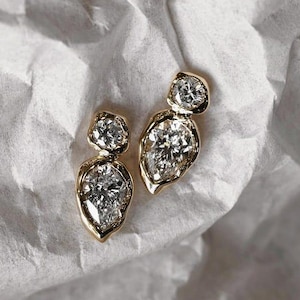 May include: A pair of gold earrings with a unique design. Each earring features a large, pear-shaped diamond and a smaller, round diamond, set in a gold bezel. The earrings are displayed on a textured white surface.