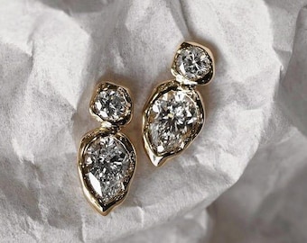 May include: A pair of gold earrings with a unique design. Each earring features a large, pear-shaped diamond and a smaller, round diamond, set in a gold bezel. The earrings are displayed on a textured white surface.