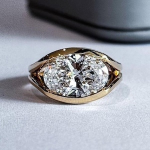 Pode incluir: Um anel de ouro com um grande diamante de corte oval. O diamante é engastado em uma moldura de bisel, e a banda é de ouro polido. O anel é exibido em uma superfície branca, com um fundo desfocado.