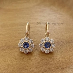May include: A pair of gold hoop earrings featuring a flower-shaped charm. Each charm has a central blue gemstone surrounded by a halo of small clear stones. The text Diamondshreejewel is visible above the earrings. The design is symmetrical.