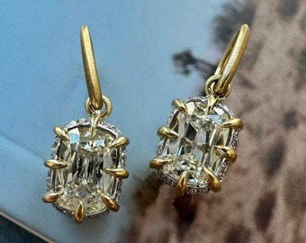 May include: A pair of gold earrings featuring large, rectangular, clear gemstones. The gemstones are set in a gold and silver frame with a gold hook. The earrings are displayed on a blue and brown patterned surface.