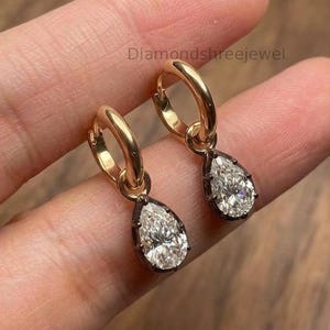 May include: A pair of gold hoop earrings featuring pear-shaped, faceted clear stones set in dark metal prongs. The earrings are marked with the text Diamondshreejewel. Each hoop has a hinged closure, and the teardrop-shaped stones hang freely from the circular gold bands.
