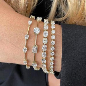 Könnte beinhalten: Vier goldene Armbänder mit funkelnden Diamantakzenten. Die Armbänder weisen verschiedene Diamantformen auf, darunter runde und Smaragdschliffe, die in zarten Goldbändern gefasst sind. Die Armbänder werden am Handgelenk vor schwarzem Hintergrund getragen.