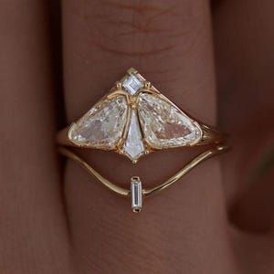 Może przedstawiać: Złoty pierścionek o unikalnym wzorze z dużym diamentem w kształcie gruszki i mniejszymi kamieniami akcentującymi. Pierścionek ma delikatną, zakrzywioną obrączkę i drugą obrączkę z prostokątnym kamieniem. Diamenty są przejrzyste i błyszczące.