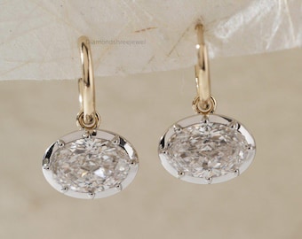 May include: A pair of gold hoop earrings with oval-shaped, clear gemstones. The gemstones are set in a silver-colored bezel with a delicate, textured design. The earrings are displayed against a neutral background.