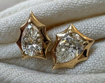May include: A pair of gold stud earrings, each featuring a pear-shaped, clear gemstone. The gemstones are set in a gold frame with a pointed, starburst design. The earrings are displayed on a textured, off-white surface.