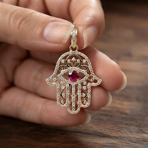May include: A gold Hamsa hand pendant featuring a central round-cut red gemstone surrounded by clear pavé-set stones. The intricate openwork design includes a bail at the top, with the text diamondshreejewel visible in the background.