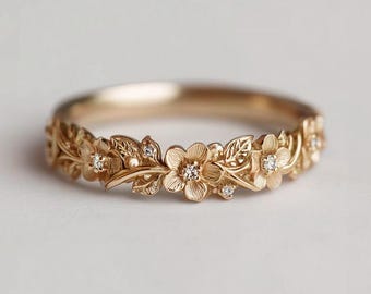 May include: A gold-colored ring with a floral design. The ring features detailed carvings of flowers and leaves, with small, sparkling gemstones set within the flower centers. The ring's band is smooth and polished, creating a contrast with the textured floral elements.