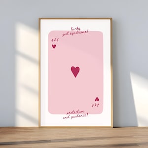 May include: A framed print featuring a pink playing card design with a large heart in the center. The card has the text "lucky girl syndrome!" at the top and "protection and guidance!" at the bottom. The card also has the number 444 and a heart symbol in the corners.