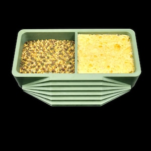 May include: A light green, two-compartment container with a textured surface. One side holds small, granular items in shades of brown, yellow, and green. The other side contains a yellow, porous sponge. The container has a decorative, layered design.