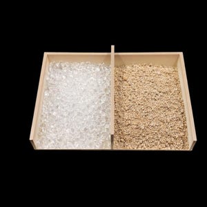 May include: A wooden tray divided into two sections, each filled with different materials. One side contains clear, crystalline stones, while the other holds small, tan, granular particles. The tray is set against a black background.