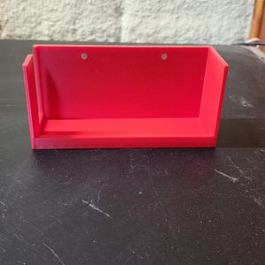 May include: A bright red, rectangular wall-mounted shelf with a flat base, vertical side panels, and a tall back panel. The back panel features two small, circular pre-drilled holes positioned horizontally near the top edge for mounting purposes.