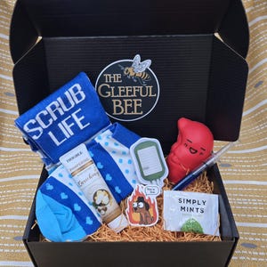May include: A black gift box filled with self-care items. Includes a blue towel with "SCRUB LIFE" text, lotion, socks, stickers, a red stress ball, pen, and mints. The box has a "The Gleeful Bee" logo.