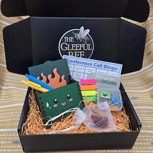 May include: A black gift box filled with shredded paper, containing a variety of office supplies. Includes a green box with a face, pens, sticky notes, a bingo card, and a small bag of red items. The box has the logo "The Gleeful Bee".