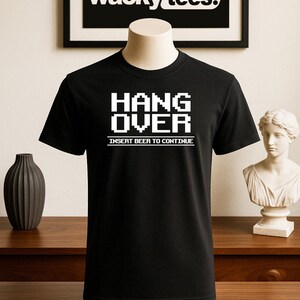 May include: Black t-shirt with the text "HANG OVER" in white, pixelated font, with "INSERT BEER TO CONTINUE" below. The shirt is displayed on a mannequin. A framed "wacky tees." logo is in the background.