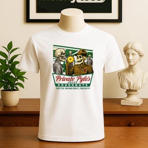 May include: White t-shirt featuring a graphic of skeleton soldiers with a donut, the text "Private Pyle's Jelly Doughnuts" and "THEY'RE PAYING FOR IT, YOU EAT IT." Above the shirt is a framed sign that says "wacky tees."