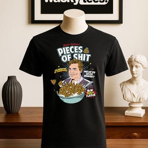 Puede incluir: Camiseta negra con un gráfico humorístico. El diseño incluye el texto "PIECES OF SHIT" y una imagen de dibujos animados de un hombre con un cuenco de cereales. La camiseta también presenta el texto "THE BREAKFAST OF CHAMPIONS" y "NEW TASTY FLAVOR".