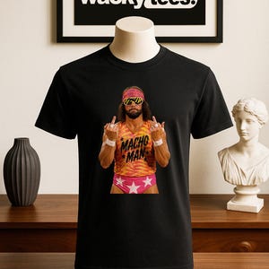 Könnte beinhalten: Schwarzes T-Shirt mit einem farbenfrohen Bild eines Wrestlers mit Sonnenbrille und Stirnband. Der Wrestler trägt ein rosa-oranges Outfit mit den Worten "Macho Man". Das Shirt hat das "wacky tees" Logo.