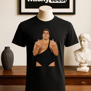 Könnte beinhalten: Schwarzes T-Shirt mit einer Grafik eines Wrestlers in einem schwarzen Trikot, der mit beiden Händen den Mittelfinger zeigt. Das Shirt wird auf einer Schaufensterpuppe präsentiert. Der Markenname "wacky tees." wird in Weiß auf schwarzem Hintergrund angezeigt.