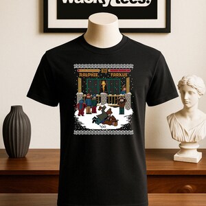 Può includere: T-shirt nero con grafica pixelata di una scena invernale con la scritta "Ralphie & Farkus". La maglietta ha un design festivo intorno allo scollo. Il logo "wacky tees." è visibile sullo sfondo.