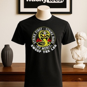 May include: Black t-shirt featuring a circular graphic with a yellow cobra, the words "Strike First, Strike Hard, No Mercy, Sweep the Leg." The shirt is displayed on a mannequin. The brand "wacky tees." is visible in the background.