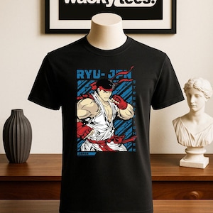 Può includere: T-shirt nera con grafica di un personaggio di un gioco di combattimento in un gi bianco, fascia rossa e guanti. Il design include il testo "RYU-JPN" e "STREET FIGHTER". La maglietta è esposta su un manichino.