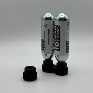 Può includere: Due cartucce di CO2 filettate argentate da 16 grammi con etichette bianche e testo nero, tenute in un supporto di plastica nero. È visibile anche un tappo di plastica nero separato. Per gonfiare i pneumatici.