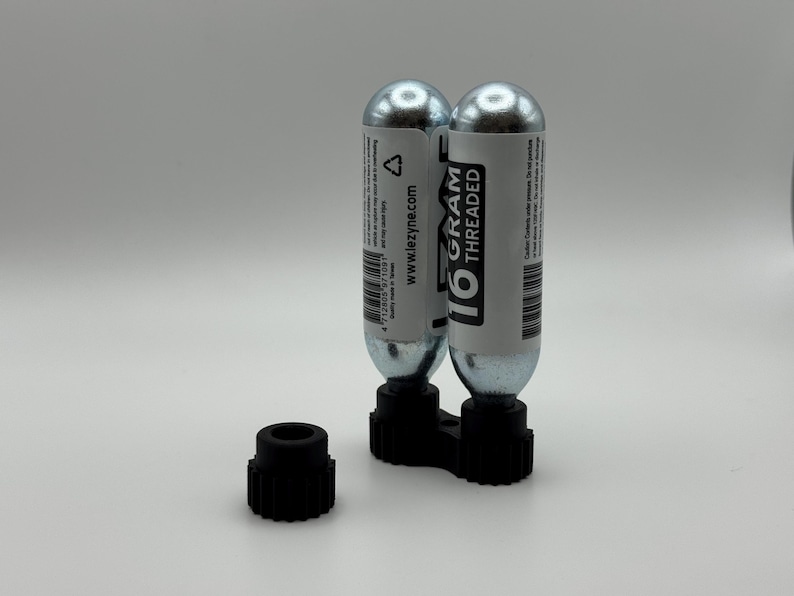 May include: Two silver 16-gram threaded CO2 cartridges with white labels and black text, held in a black plastic mount. A separate black plastic cap is also visible. The cartridges are for inflating tires.