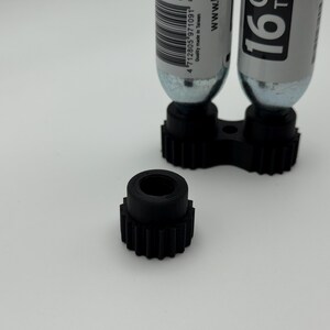 May include: A black, 3D-printed gear-shaped component with a central hole, positioned in front of a black holder with two silver CO2 cartridges. The cartridges have white labels with black text. The background is plain white.
