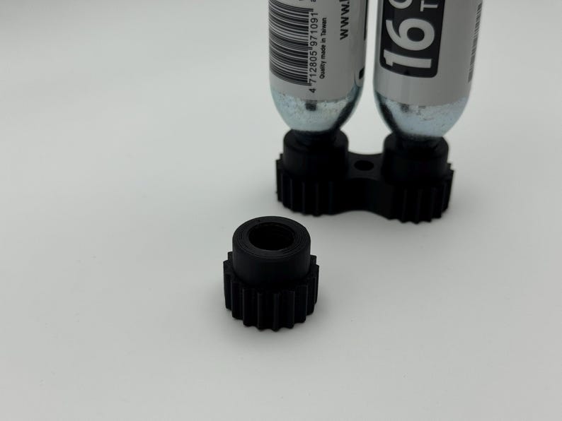 May include: A black, 3D-printed gear-shaped component with a central hole, positioned in front of a black holder with two silver CO2 cartridges. The cartridges have white labels with black text. The background is plain white.
