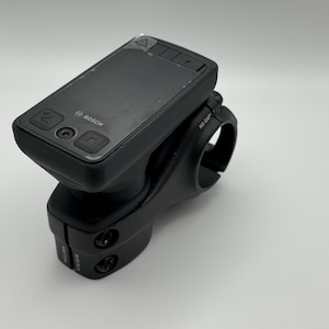 Puede incluir: Un ordenador de bicicleta negro con el logotipo de Bosch. El dispositivo tiene una pantalla rectangular y varios botones. Está montado en una potencia negra con una abrazadera para fijarlo al manillar de una bicicleta.
