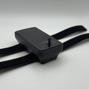 Puede incluir: Un dispositivo electrónico negro con botones y la marca "BOSCH" está sujeto a dos correas de tela negras. El dispositivo tiene un tornillo en la parte superior y está sobre un fondo blanco.