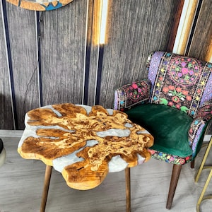 Può includere: Un tavolino in legno unico, a forma libera, con un riempimento in resina colorata. Il tavolo ha tre gambe in legno scuro. Il piano del tavolo presenta un motivo a venature naturali con resina grigio chiaro. Il tavolo è lungo circa 76 cm.