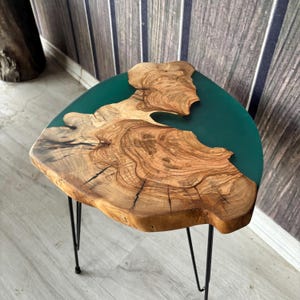 May include: A handcrafted side table with a live edge wooden top and teal epoxy resin. The table has a natural wood grain and sits on three black metal hairpin legs. The table is approximately 61 cm wide.