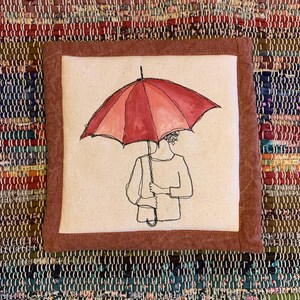 May include: Square fabric art piece featuring a person holding a red umbrella. The umbrella is open, with a gradient of red hues. The artwork is framed with a brown border and set against a colorful woven background.