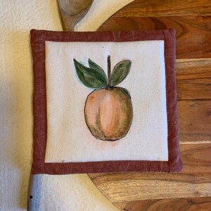 May include: A square, off-white pot holder with a watercolor-style peach design. The peach is surrounded by green leaves and a brown stem. The border is a dark red fabric. A wooden spoon is placed above the pot holder.