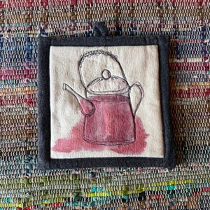 May include: A square fabric potholder featuring a cream-coloured centre panel with a black embroidered outline of a teapot. The teapot is filled with a watercolour-style red pigment. The potholder has a dark grey fabric border and a small hanging loop.