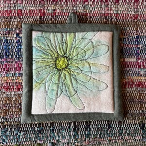 May include: A square fabric potholder featuring a central light green and pale blue flower design with dark thread outlines. The flower has a bright yellow center and is framed by a solid dark olive green border with a small fabric hanging loop.