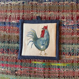 May include: Square, quilted pot holder with a blue denim border and hanging loop. The center features a watercolor-style illustration of a blue rooster with a red comb and wattle. The background is off-white.