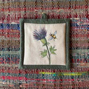 Puede incluir: Salvamanteles cuadrado de color blanquecino con un diseño de acuarela de una flor de cardo morada y una abeja amarilla y negra. El salvamanteles tiene un borde verde y un lazo para colgar.