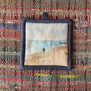 Puede incluir: Salvamanteles cuadrado y acolchado con una escena de playa. La obra de arte representa dos figuras en una playa de arena, con un océano y un cielo azul claro. El salvamanteles tiene un borde azul oscuro y un lazo para colgar.