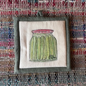 May include: A square fabric potholder with a dark olive green border and a cream-colored center panel. The center features a detailed illustration of a glass jar filled with green pickles, topped with a pink lid and a small green sprig.