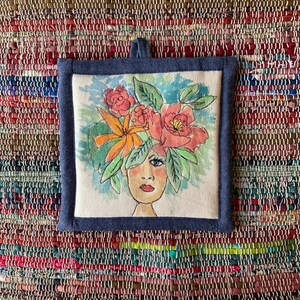 Pode incluir: Pega-panela quadrada, costurada à mão, com um retrato floral. A obra de arte apresenta o rosto de uma mulher com uma vibrante coroa de flores em tons de rosa, laranja e verde. A pega-panela é emoldurada com tecido de ganga azul escuro e tem uma argola para pendurar.