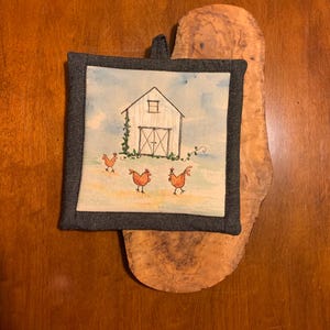 May include: Square pot holder with a watercolor painting of a white barn with a black outline, a blue sky, and three chickens. The pot holder has a dark gray border and a loop for hanging.