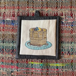 May include: Square, off-white pot holder with a dark gray border and hanging loop. The center features an embroidered stack of pancakes with syrup, blueberries, and a pat of butter on a blue plate.