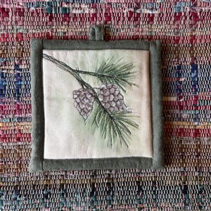 May include: A square fabric potholder featuring a central panel with an embroidered design of two pinecones on a branch with green needles. The central panel is off-white and is framed by a solid olive green fabric border with a hanging loop.